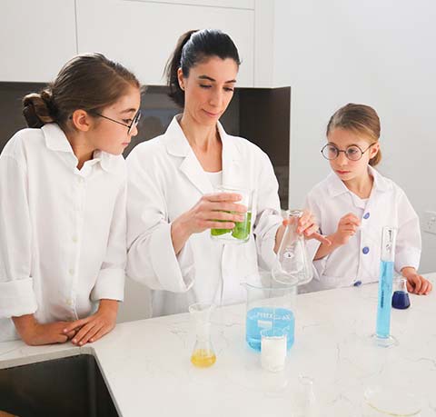 Nadine and two daughters wearing lab coats pouring liquid into scientific glassware in chemistry lab